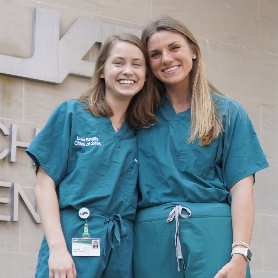 Two girls wearing green scurbs smiling for photo with the UAB School of Dentistry building sign in the background